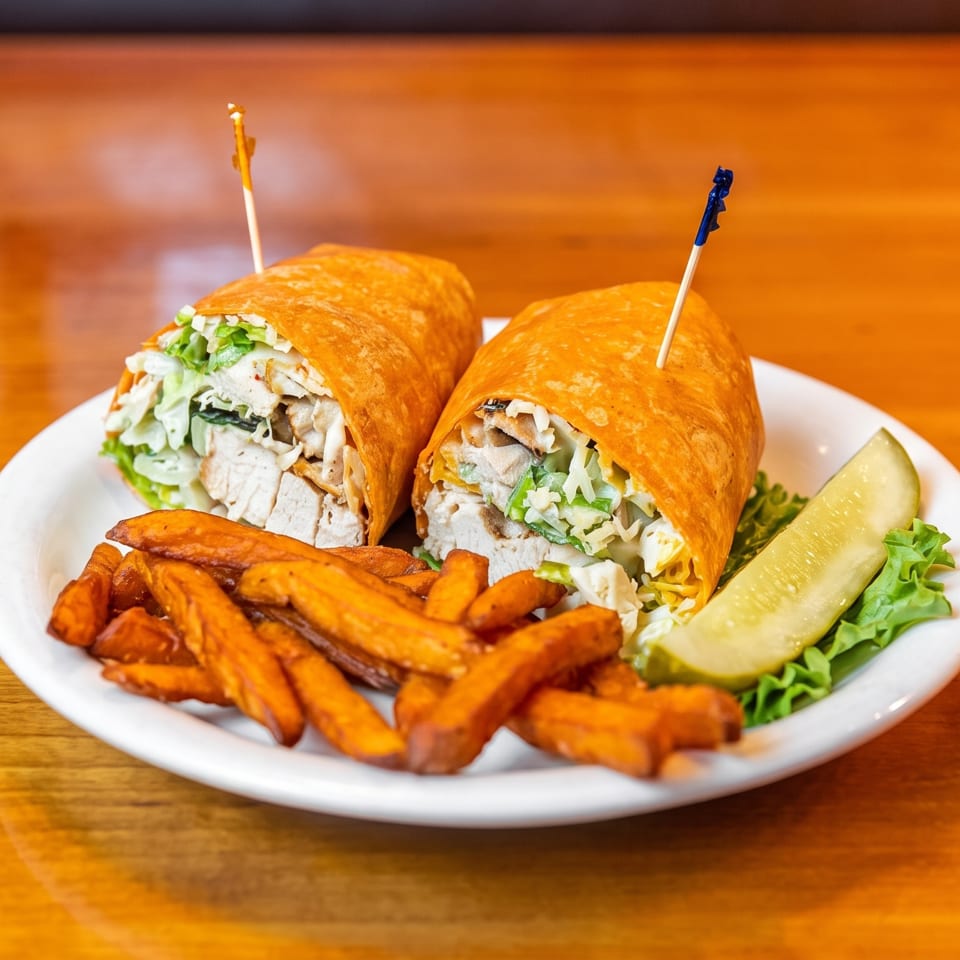 Two chicken wrap halves with lettuce and dressing, sweet potato fries, and a pickle spear on a white plate.