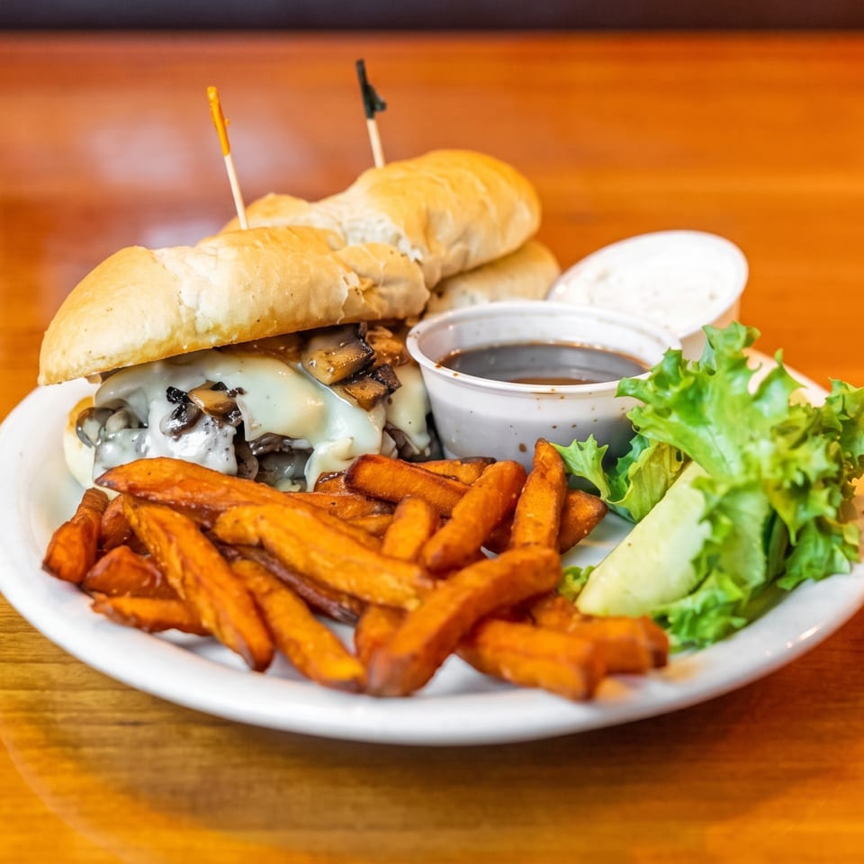 Cheesesteak sandwich with mushrooms, sweet potato fries, salad, and dipping sauces on a white plate.